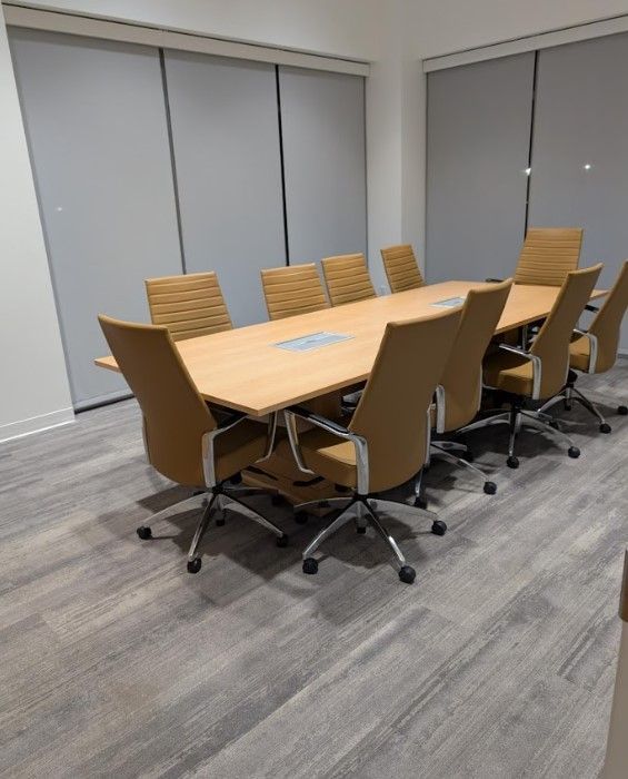 A conference room with a long table, tan chairs, gray shades, and carpet.