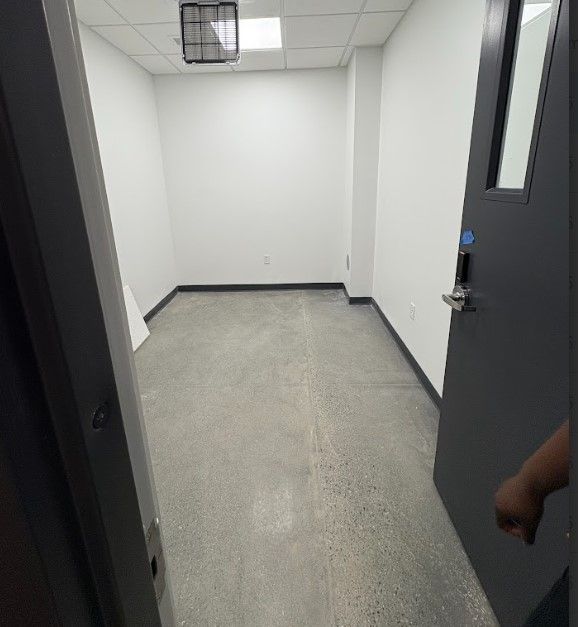 A small, empty room with white walls and a gray floor. A door is open on the right.