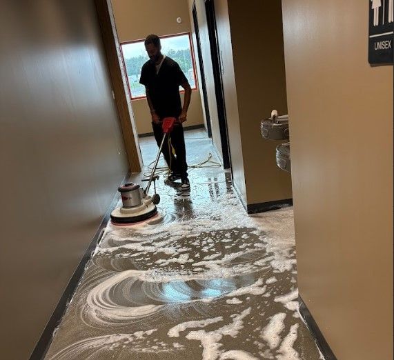 A person scrubs a hallway floor with a machine; soapy water is on the floor.
