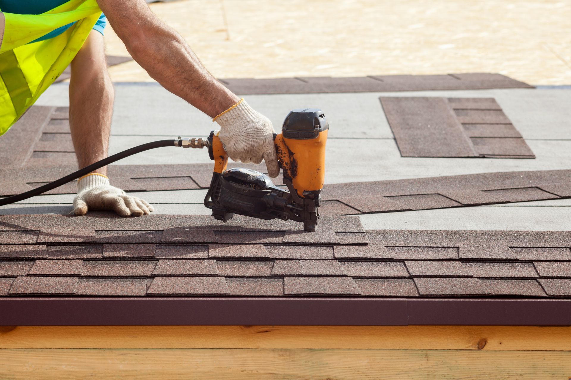 Worker using a nail gun to fasten asphalt shingles on a roof during installation.