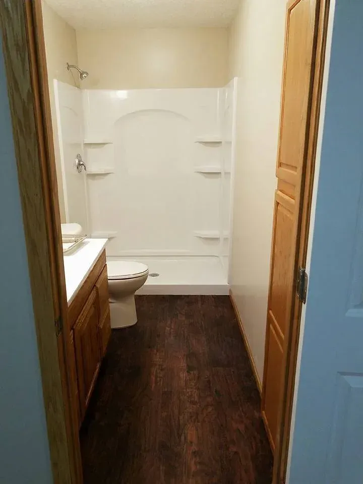 Small bathroom with wood-look flooring, white shower/tub combo, toilet, vanity and closed wooden door.
