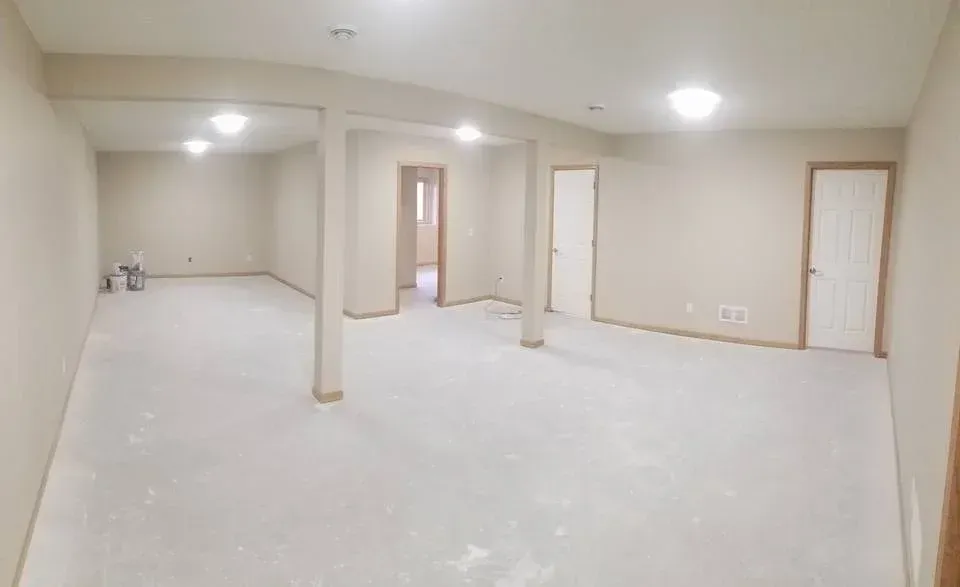 Finished basement with neutral walls and floors, several doors, support beams, and overhead lights.
