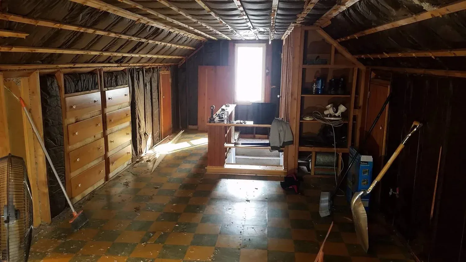 Interior of a dilapidated room with exposed wooden beams, plywood, and a small window. Shovels and tools scattered about.