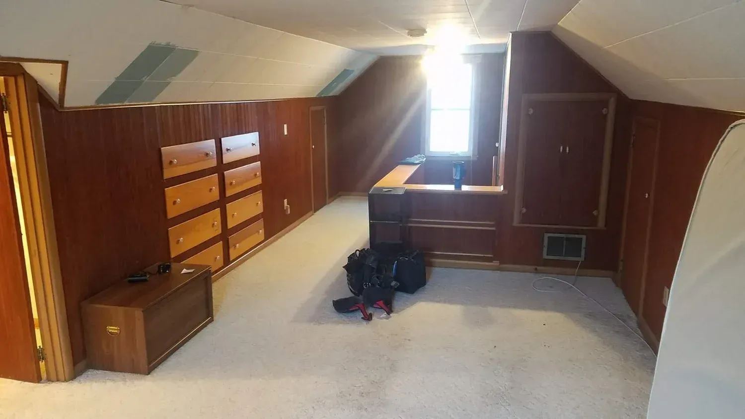 Attic bedroom with wood paneling, multiple dressers, and a small window letting in light.