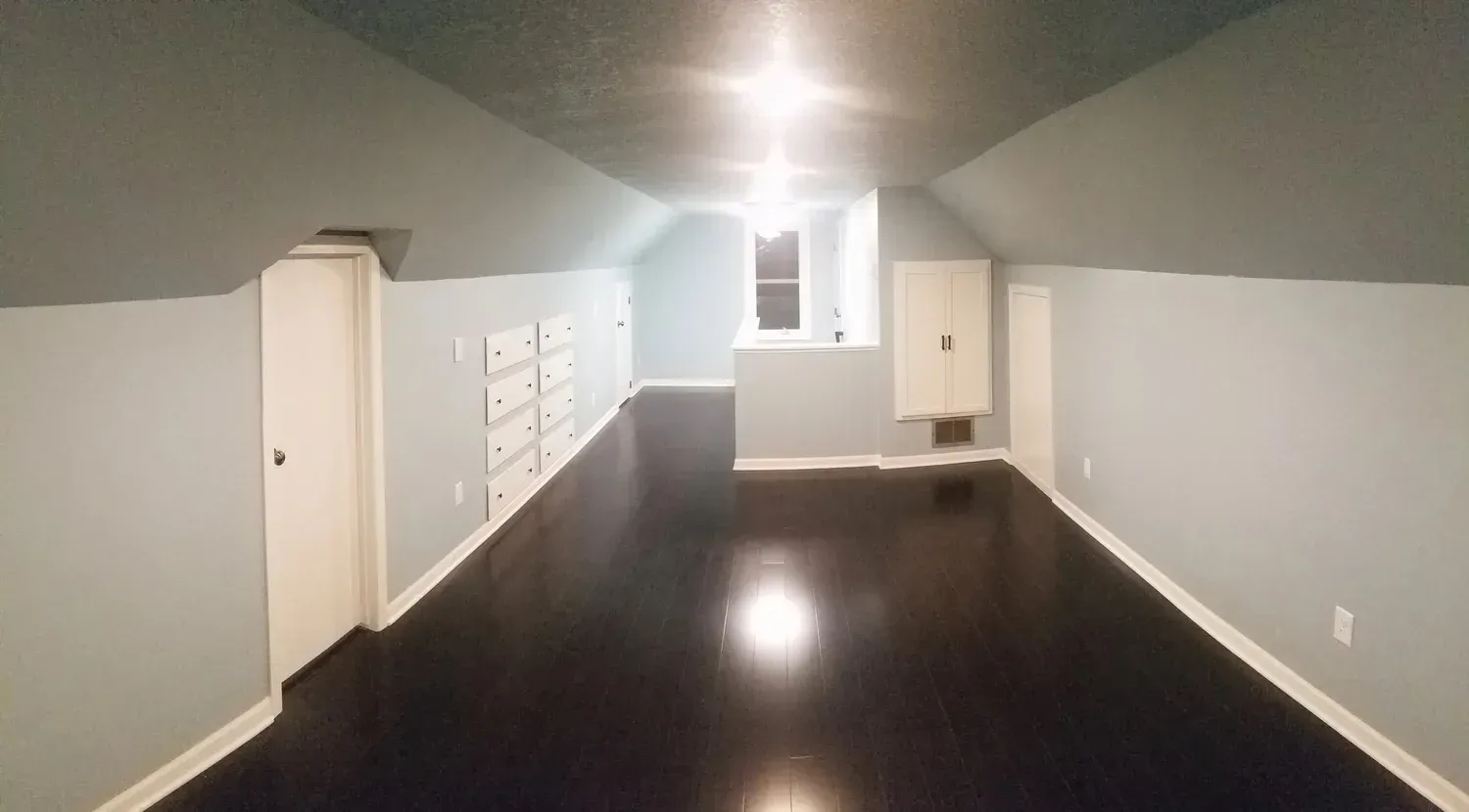 A long, narrow attic room with dark wood floor, light blue walls, and white storage.