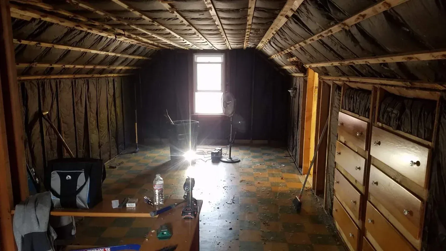 An unfinished attic with dark walls, a window, and a dresser. Sunlight streams in, illuminating dust.