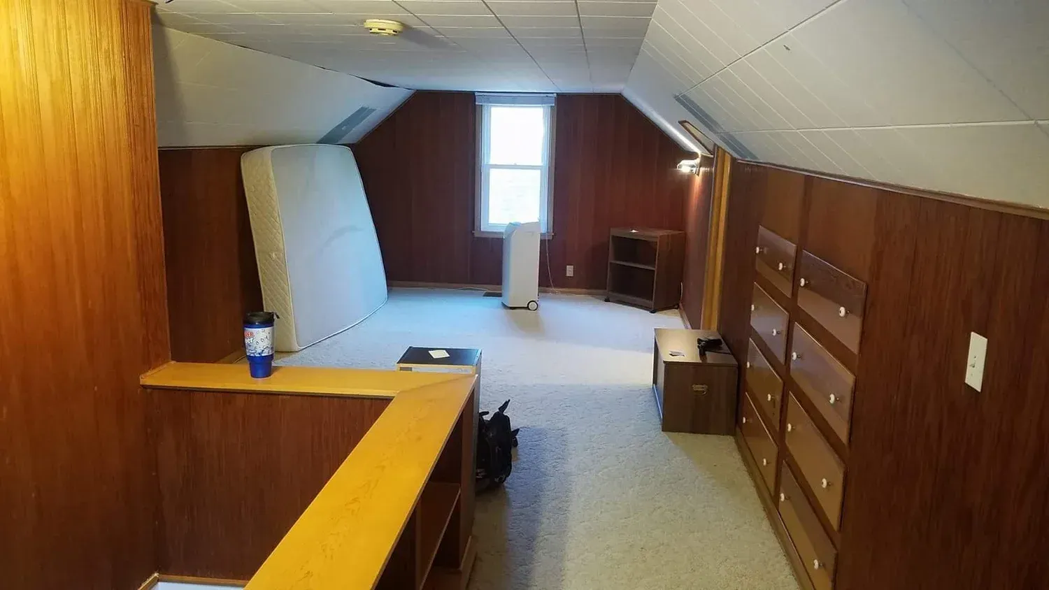 Attic room with wood paneling, a mattress, built-in drawers, and a small window.