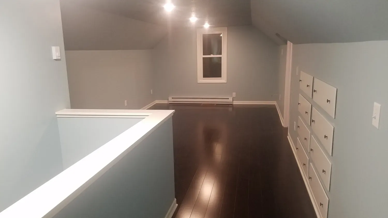 Light blue room with dark wood floor, white railing and cabinets, window at the end.
