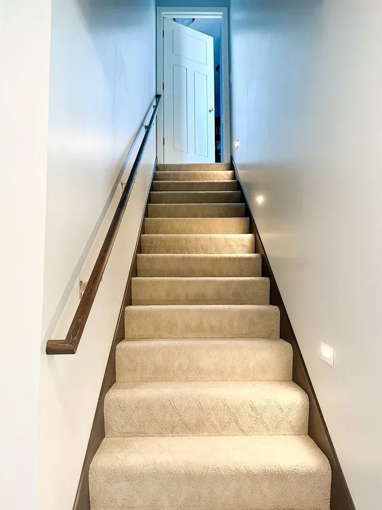 Staircase with beige carpet leading up to a white door; a wooden handrail runs along one side.