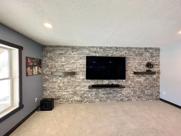 Living room with grey brick accent wall, mounted TV, floating shelves, and light carpet.