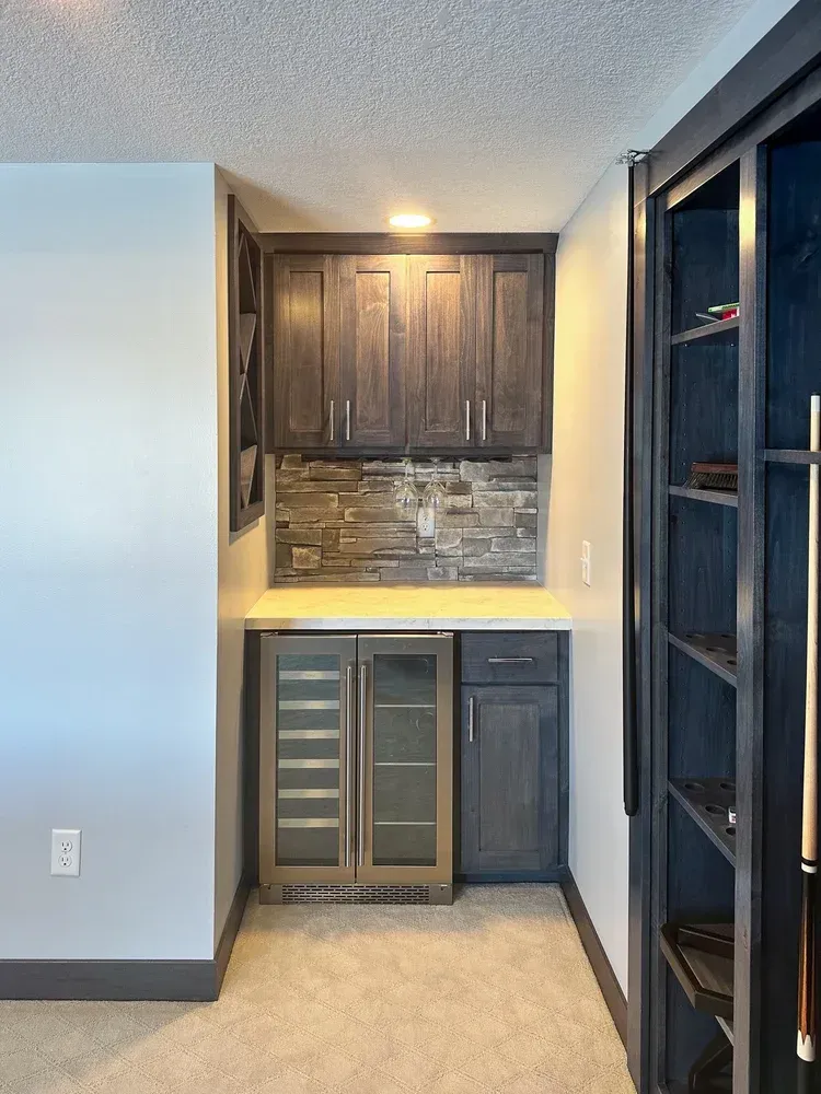 Dark gray home bar with wine fridge, cabinets, stone backsplash, and shelves.