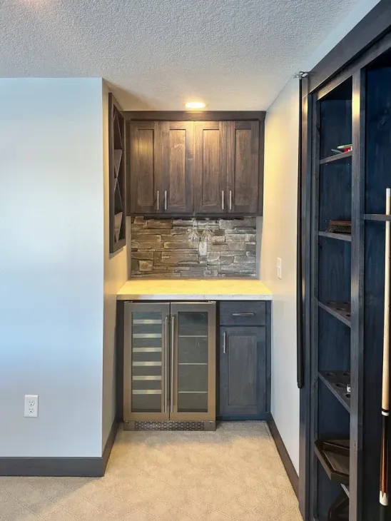 A small bar area with gray cabinets, a wine fridge, and a stone backsplash.