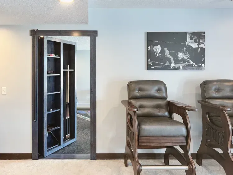 Hidden room door in dark wood frame opens to reveal shelves holding sports equipment, next to bar stools.