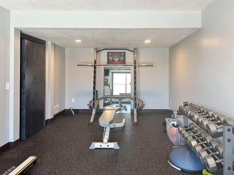 Home gym with weight rack, bench, dumbbells, and a mirror. Black flooring and gray walls.