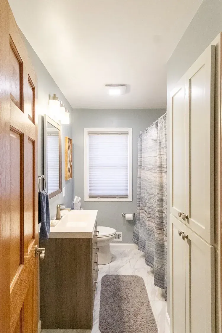 Narrow bathroom with light blue walls, white cabinets, and a gray patterned shower curtain.