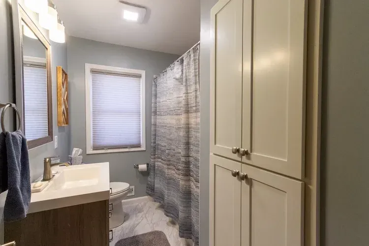 Bathroom with white vanity, toilet, and tall storage cabinet. Gray walls, patterned shower curtain, window with blinds.