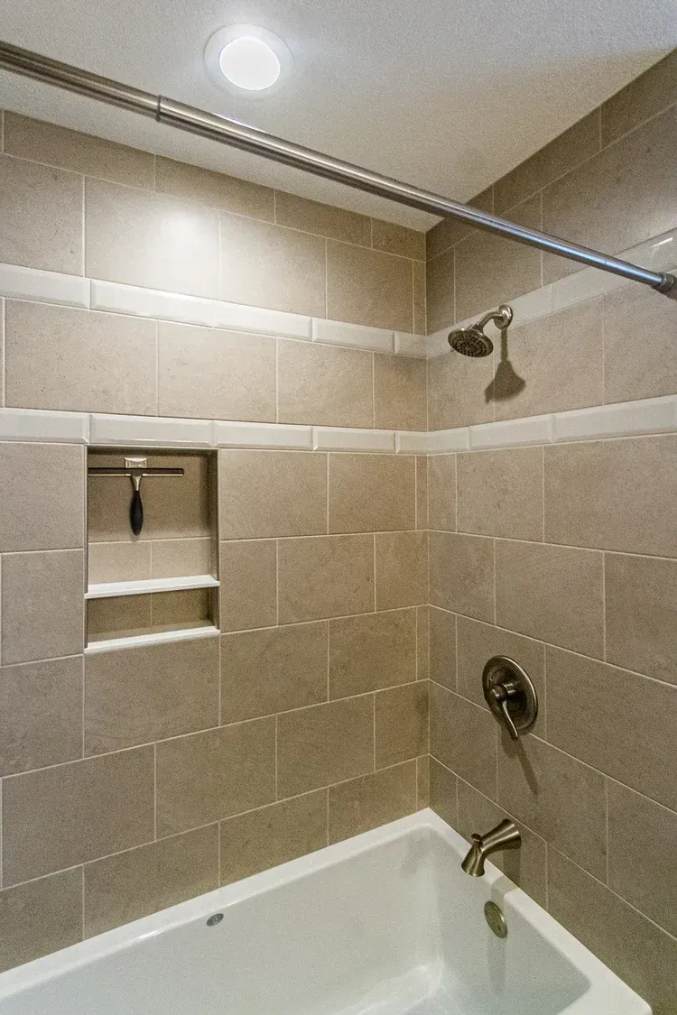 Tiled shower stall with a built-in shelf, a showerhead, and a bathtub. Beige tiles with a white accent stripe.