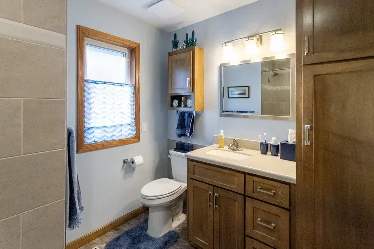 Bathroom with toilet, vanity, mirror, and shower. Neutral walls, wooden cabinets, blue accents.