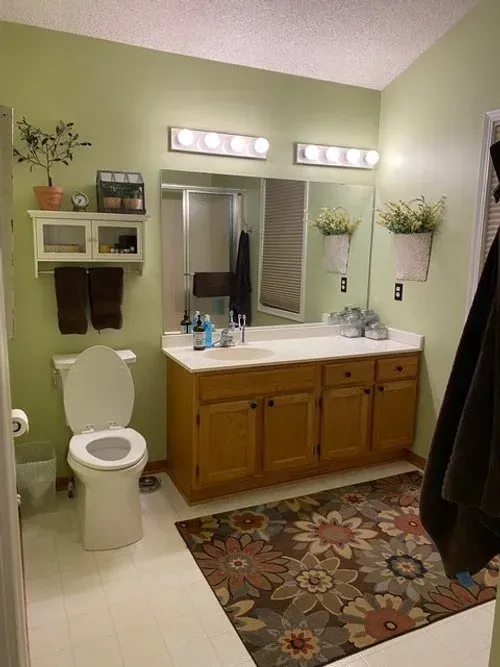 Bathroom with light green walls, wooden vanity, floral rug, toilet, and decorative plants.