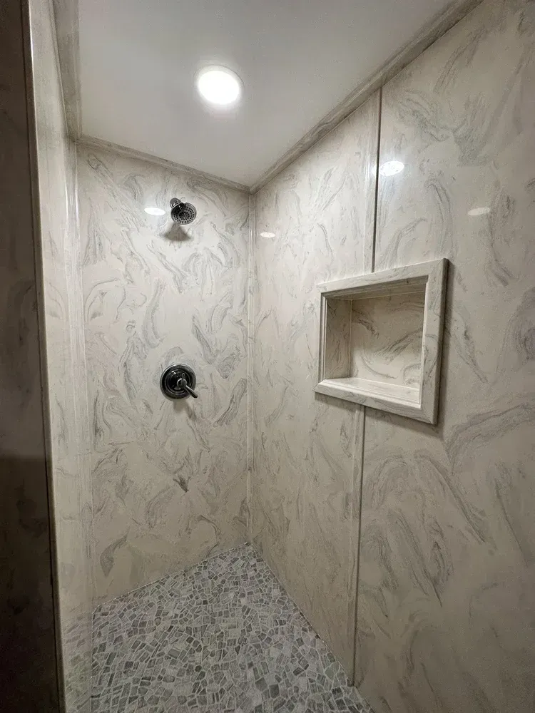 Shower stall with marbled white and gray walls, a built-in shelf, and pebble tile floor.