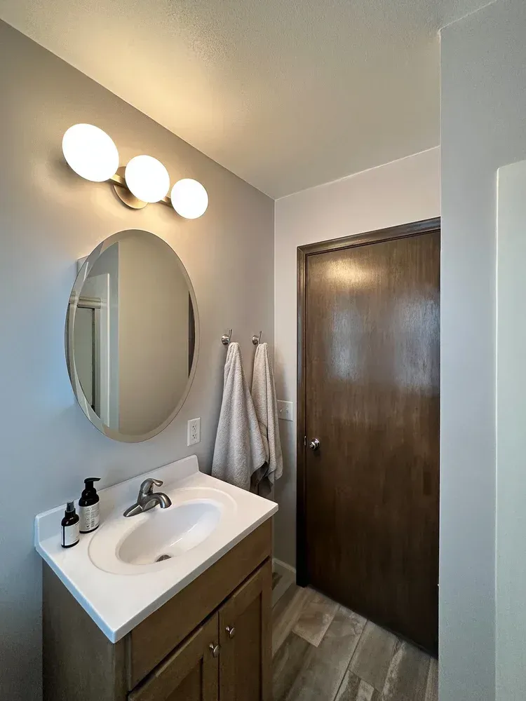Bathroom with vanity, oval mirror, globe light, and dark wooden door. Gray walls, white sink and towels.