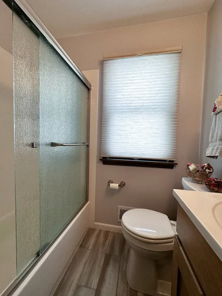 Bathroom with frosted glass shower doors, toilet, window with blinds, and vanity.