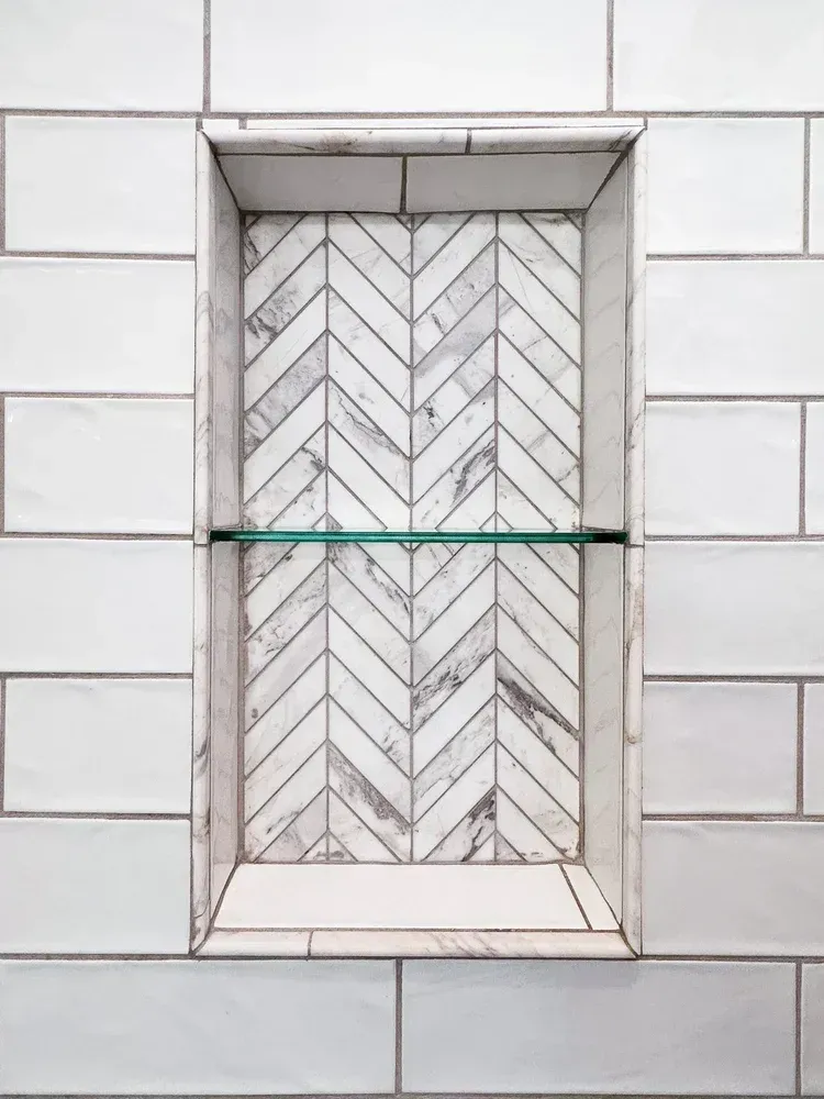 White tiled shower niche with herringbone-patterned marble backing, glass shelf.