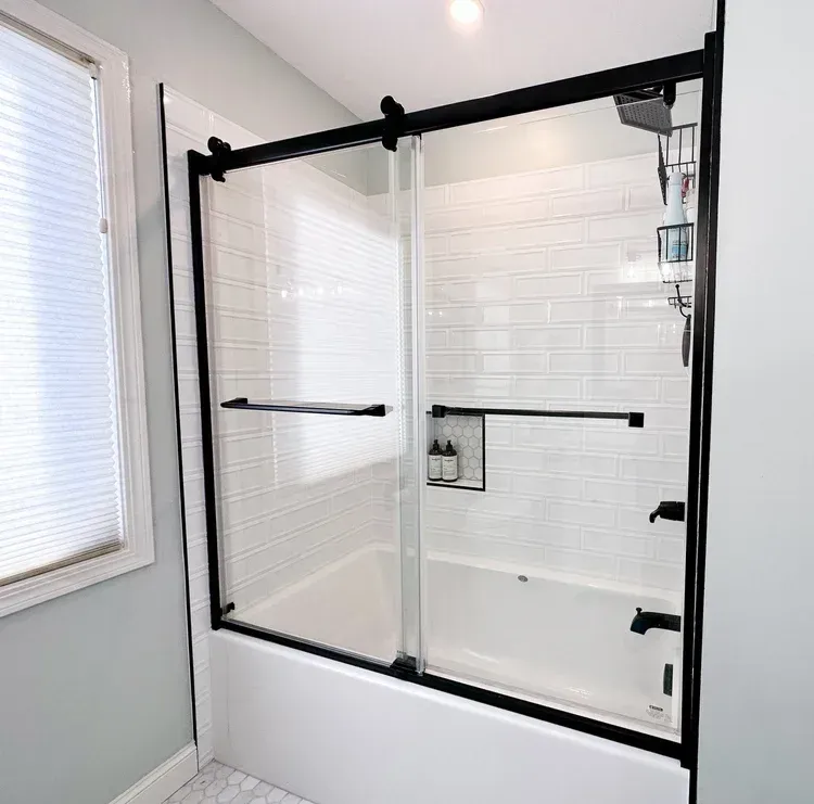 Black framed sliding glass shower doors on a white tub, white tiled walls, gray walls, and a window.