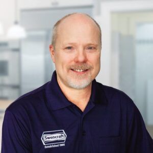 Middle-aged man with a light beard, wearing a dark blue Swencraft polo shirt, smiling indoors.