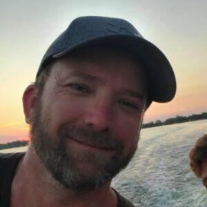Man with a beard wearing a baseball cap smiles on a boat, sunset in the background.