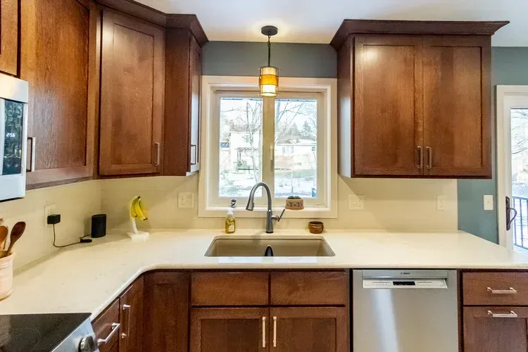 Kitchen with brown cabinets, white countertop, stainless steel sink and faucet, and a window with a pendant light.