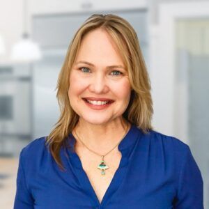 Blond woman smiling, wearing a blue shirt and necklace, in an office setting.