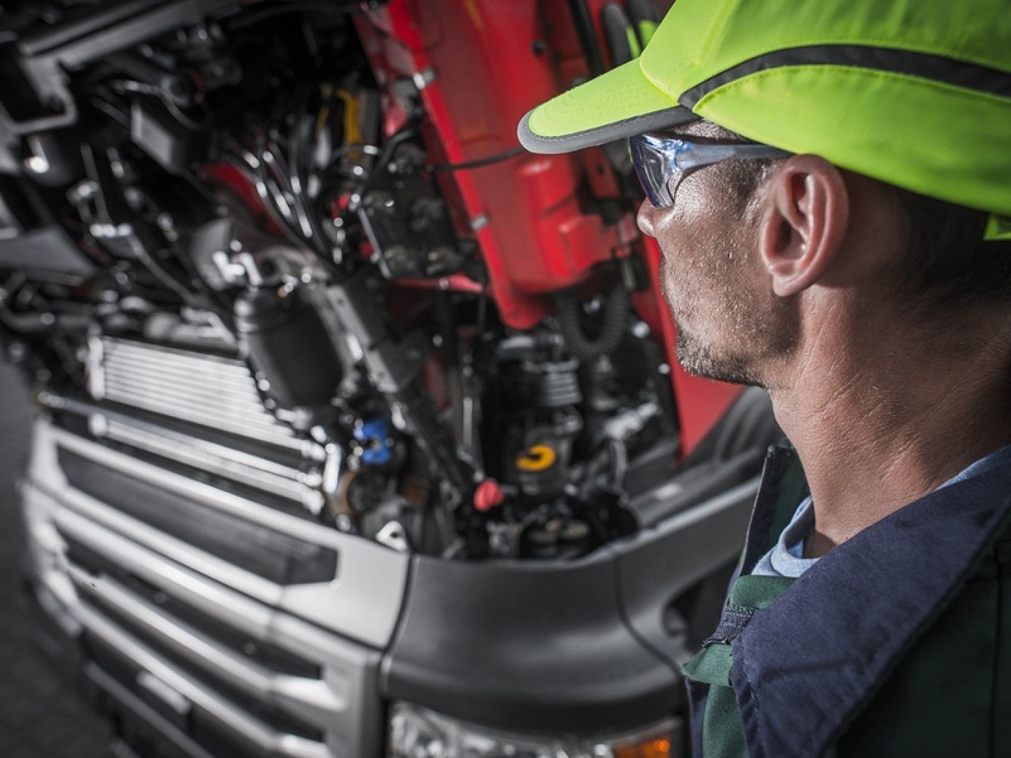 A man wearing a hard hat and safety glasses is looking under the hood of a truck.