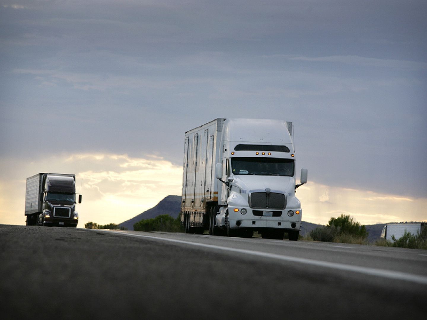 Two semi trucks are driving down a highway at sunset.