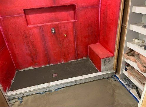 Red-walled shower stall under construction with a built-in bench and niche. Grey mortar covers the floor.