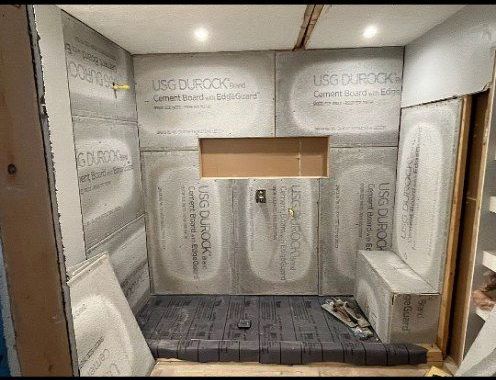 Shower area under construction, lined with gray cement board. A built-in bench and niche are visible.