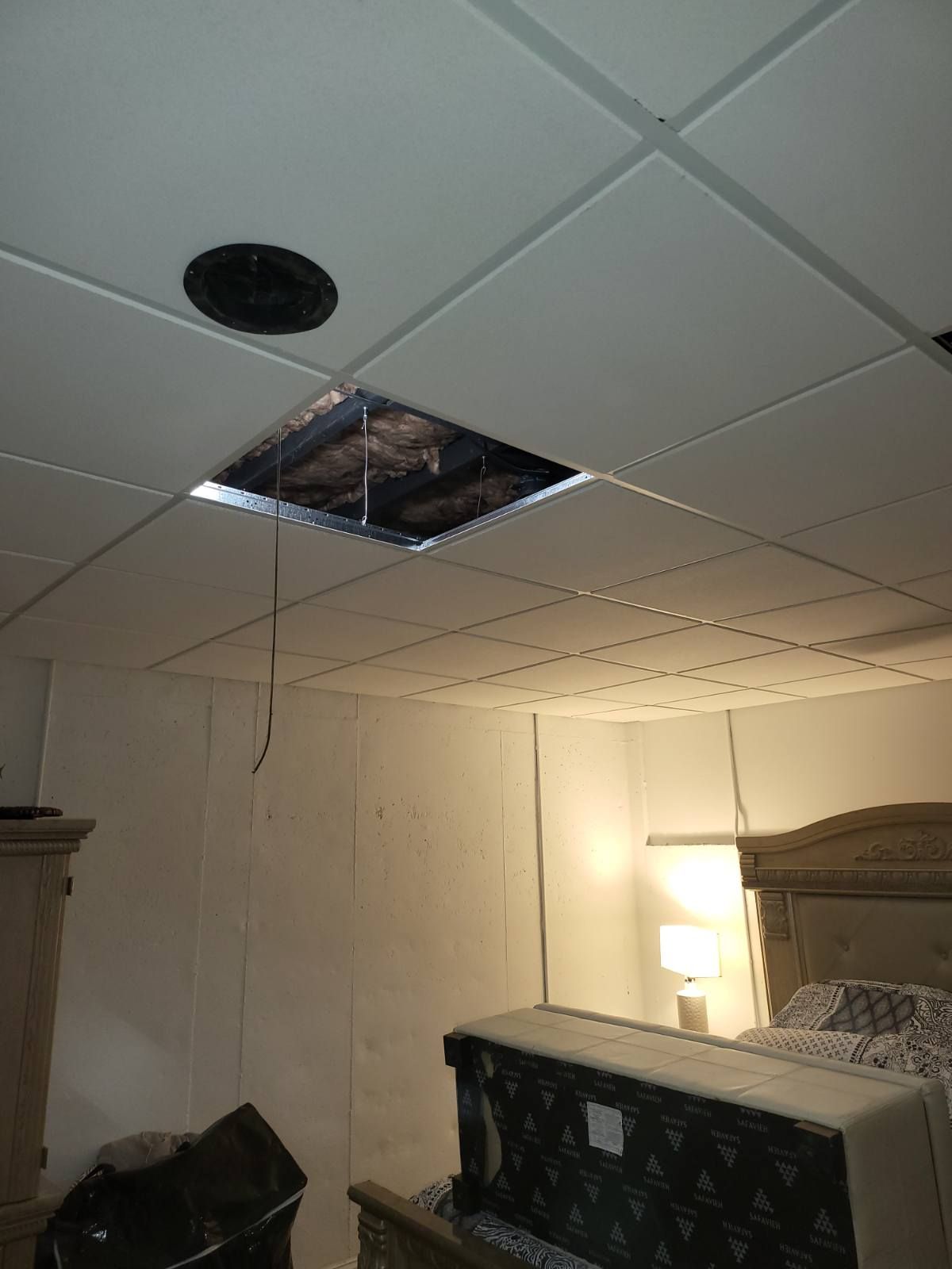 A ceiling tile is missing, revealing a hole in the ceiling of a bedroom. Debris and wires hang down.