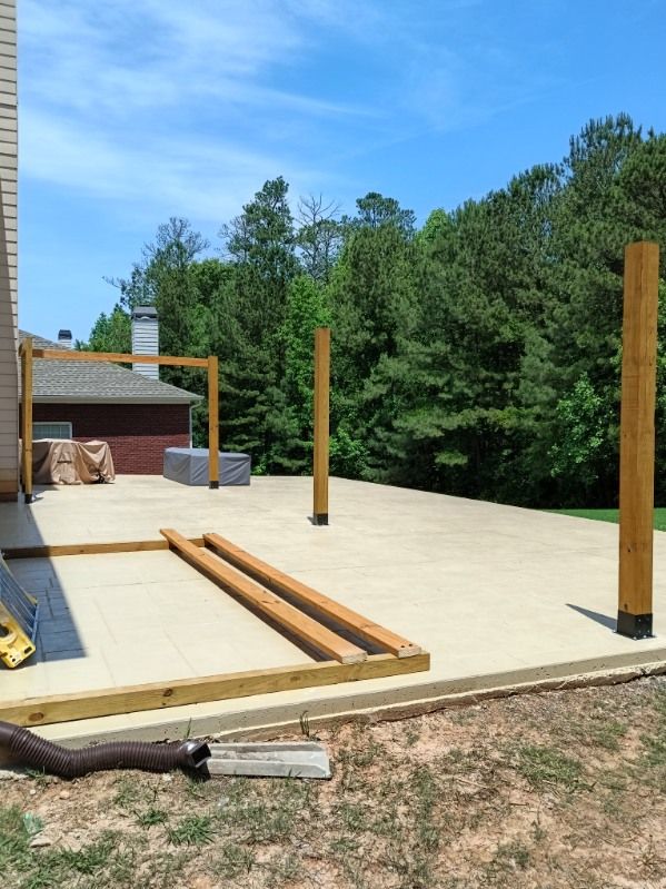 Construction of a wooden pergola on a concrete patio. Two upright support beams are in place, with others lying on the ground. The setting is outdoors on a sunny day.