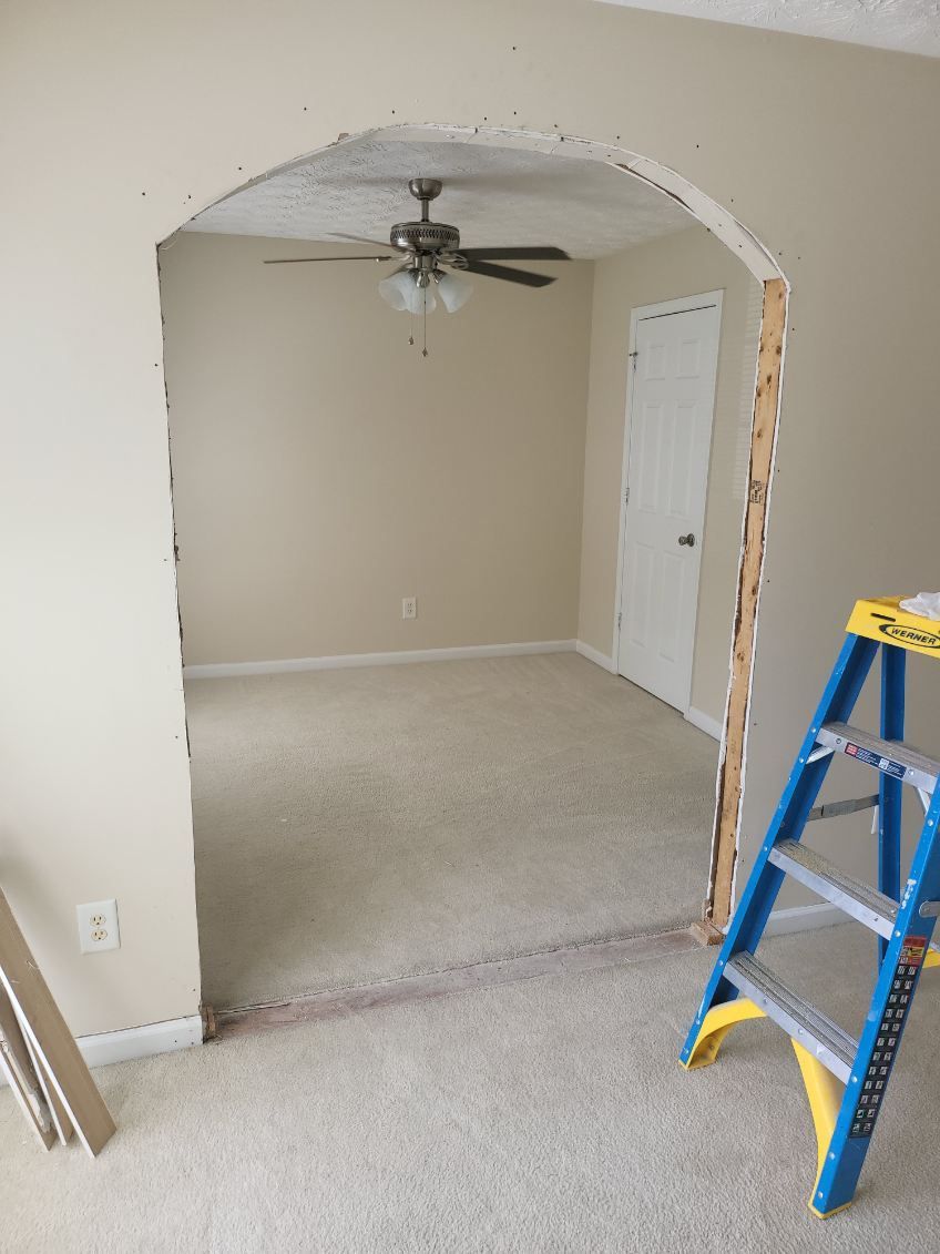 A doorway archway under construction, revealing a room with carpet and a ceiling fan. A ladder is next to the opening.