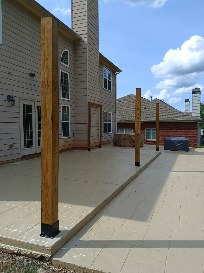 Exterior view of a beige-colored concrete patio with several wooden posts erected, set against a two-story beige house with a chimney.