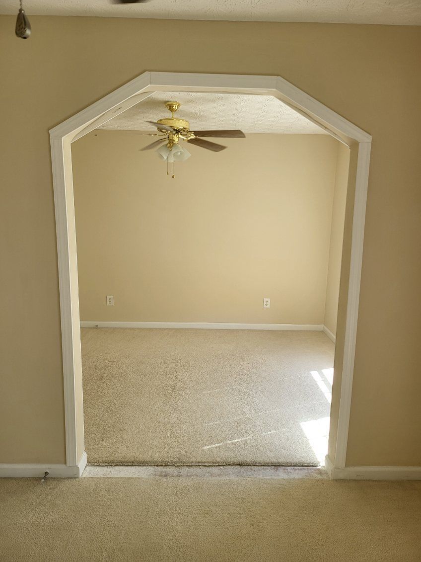 A view through an arched doorway into a room with beige walls and a ceiling fan. The room has carpeted flooring.