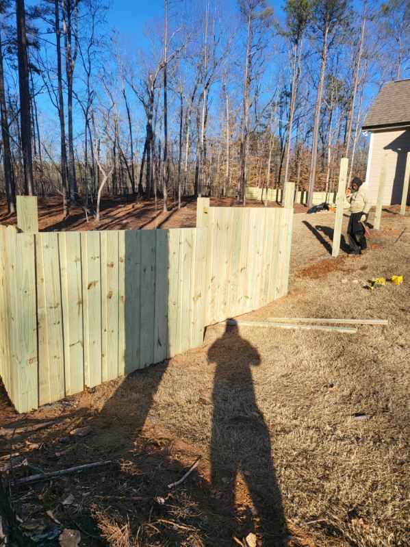 A curved wooden fence being built outdoors. A person works on the fence on a sunny day, with trees and a house in the background.