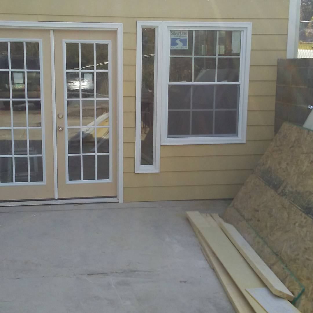 A yellow-sided building with a patio, French doors, a window, and wood siding. Piles of wood and plywood boards lay on the side.