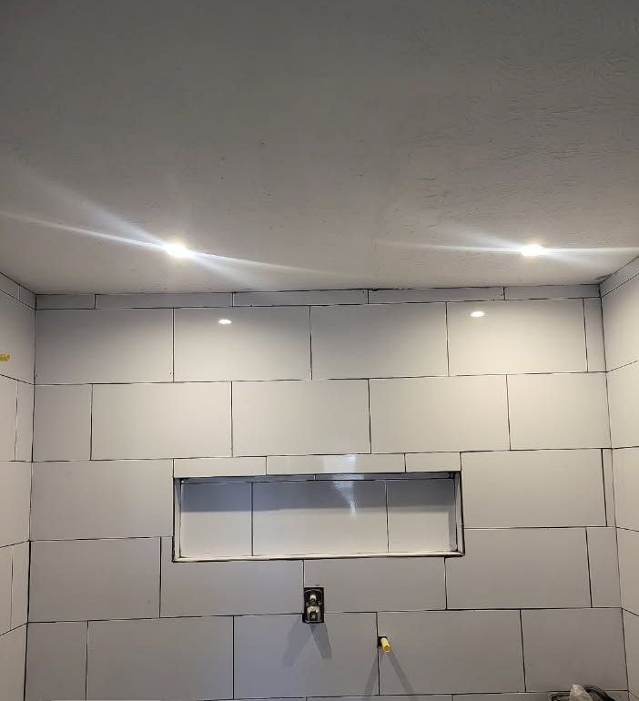 Bathroom wall in progress with white tiles and recessed shelf. Recessed lights are installed on the ceiling.