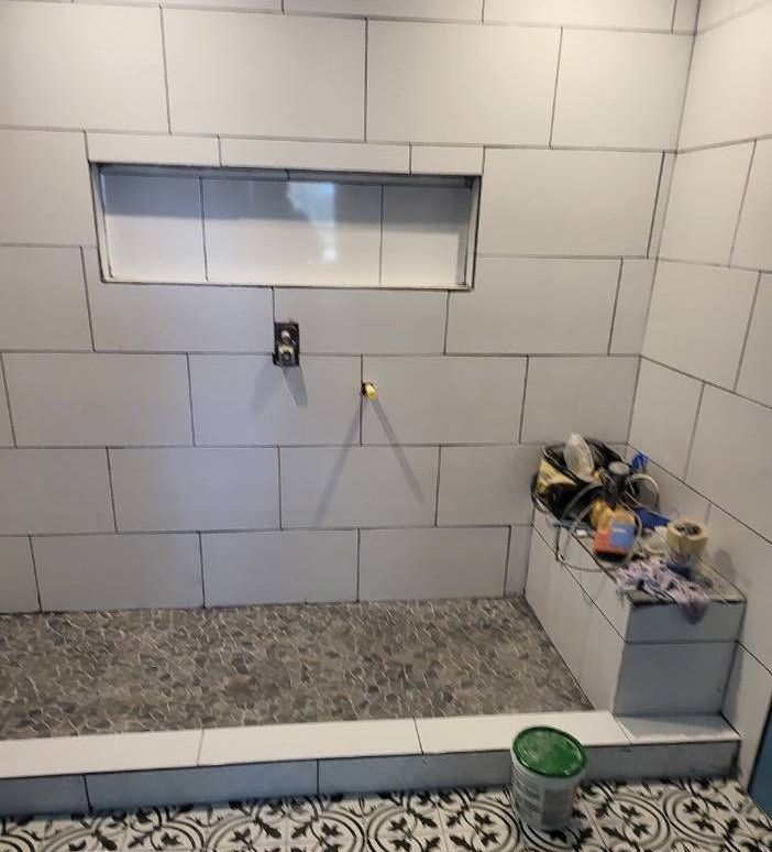 Bathroom shower with white tiled walls and a gray pebble floor. There's a built-in shelf and a step-up bench.