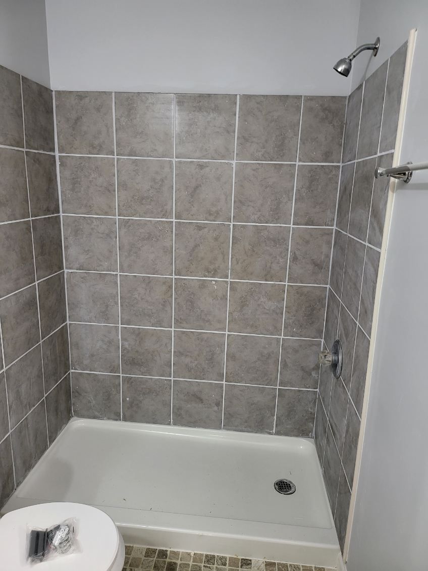 Shower stall with gray tiled walls and a white base. Silver shower fixtures and a towel rack are visible.