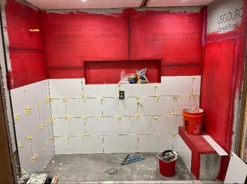 A shower under construction, featuring white tiles being laid on a red waterproof backing. A red bench and recessed shelf are also present.