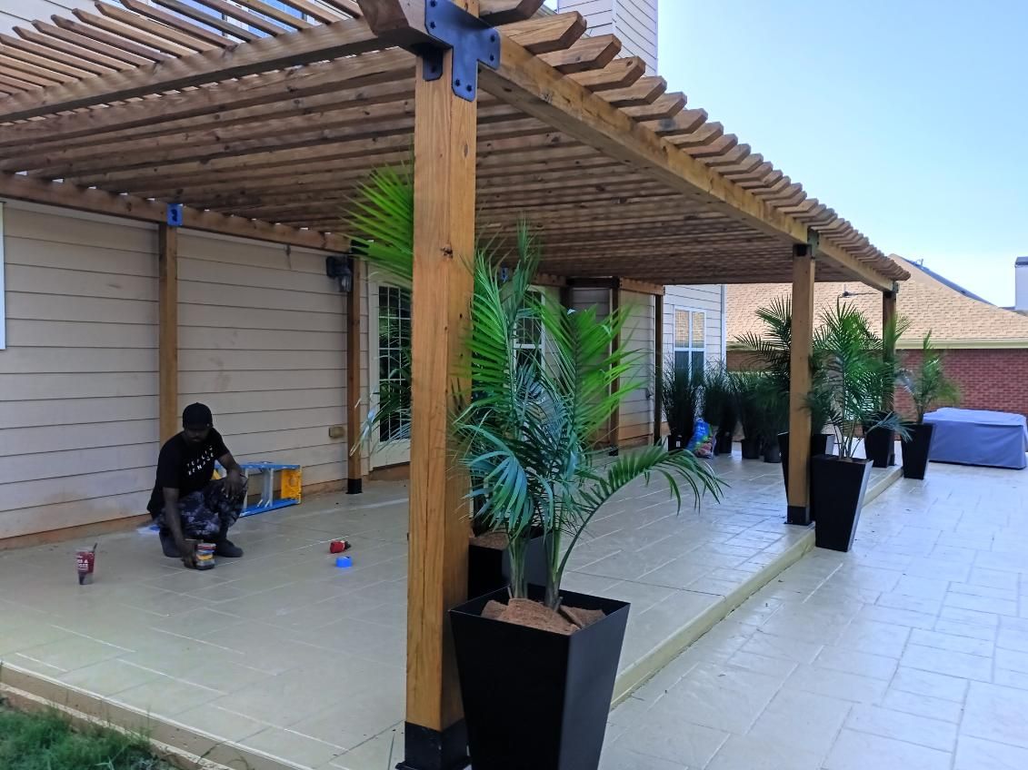 A person paints a light-colored patio under a wooden pergola with potted plants against a beige house.