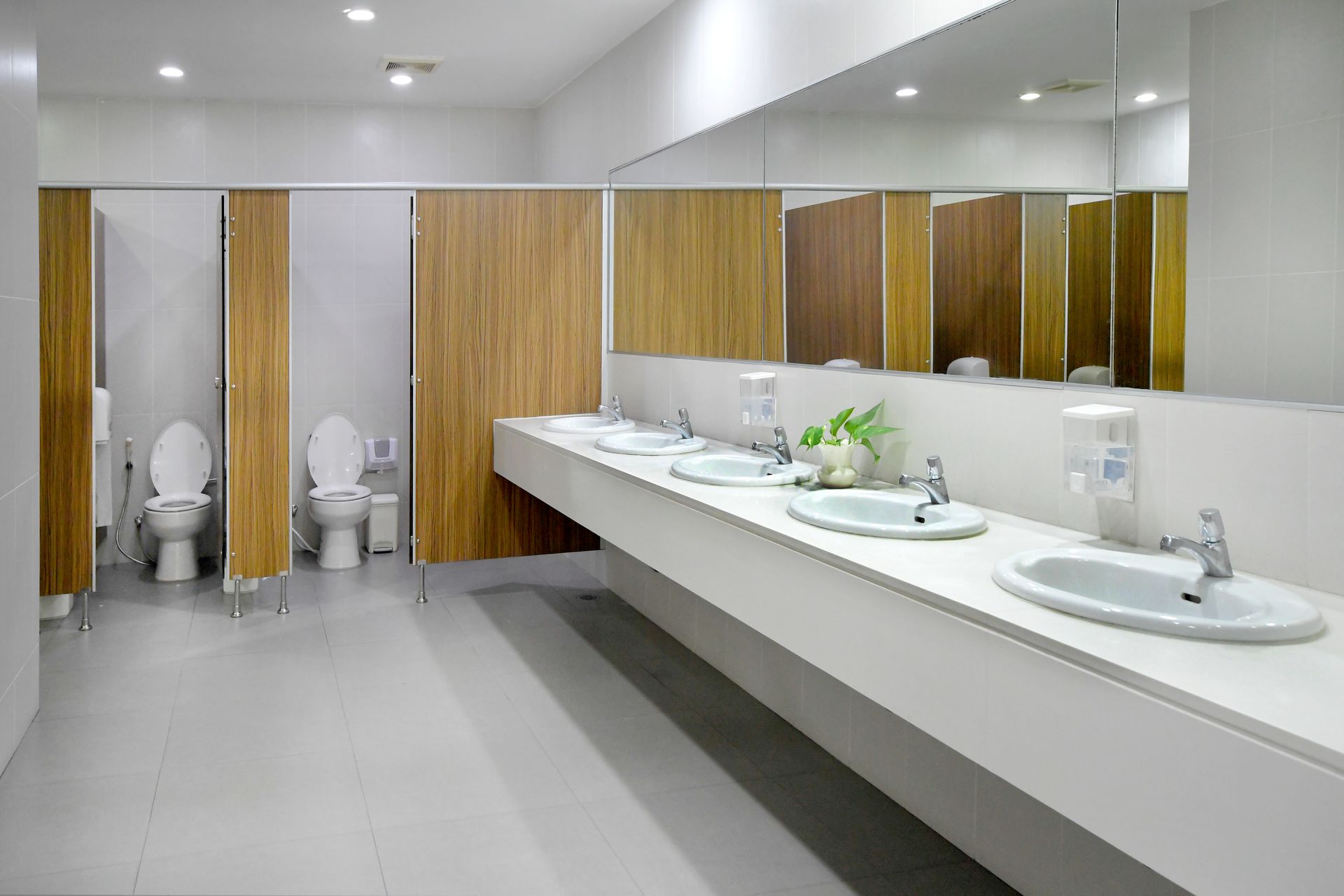 Public restroom with white sinks and countertop, wooden stalls, and large mirrors. Fluorescent lights illuminate the space.