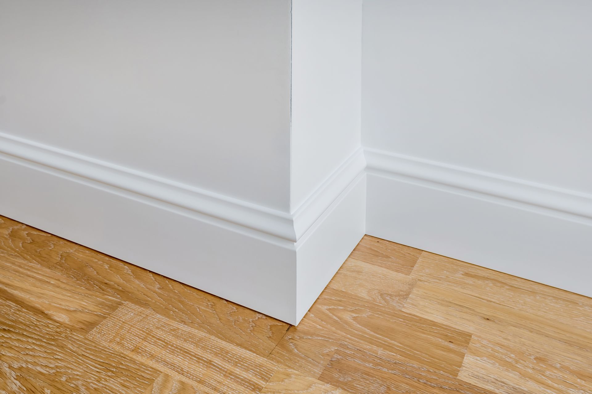 Corner of a room with white walls, trim, and light wood-colored parquet flooring.